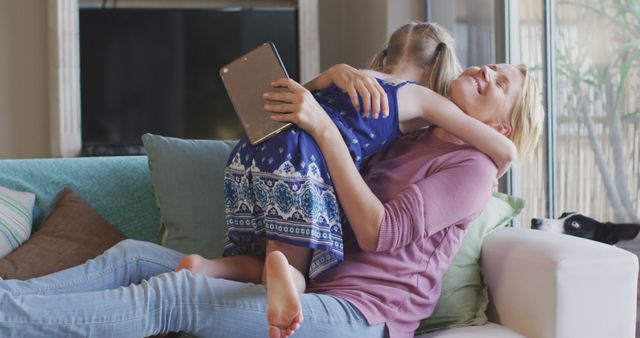 Happy caucasian daughter embracing mother holding tablet lying on sofa at home. Motherhood, childhood, love, togetherness and domestic life.