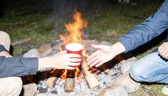 Passing red cup by campfire while friends warm hands around bonfire at night
