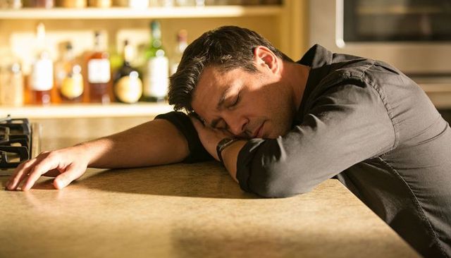 Exhausted man resting on kitchen counter, leaning head, wristwatch, moody home bar scene