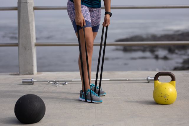 Front view low section of a strong Caucasian woman wearing sportswear exercising outdoors by the seaside on a sunny day, strength training holding black rubber tape with her foot and her hands, kettlebell and ball next to her..