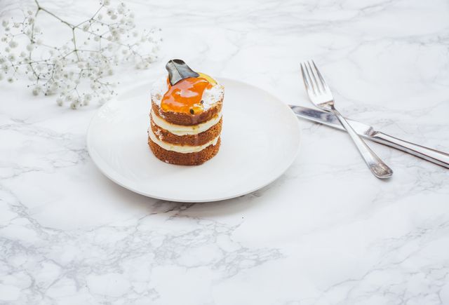 Sophisticated small cake served on white plate with an orange glaze topping, paired with a knife and fork next to it on marble table. Perfect for use in culinary blogs, dessert recipe websites, fine dining promotions, and elegant food presentation displays.