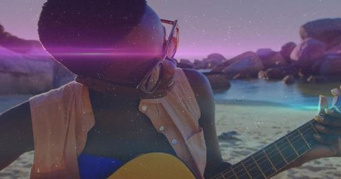 Strumming woman wearing sunglasses on sunset beach with acoustic guitar and lens flare