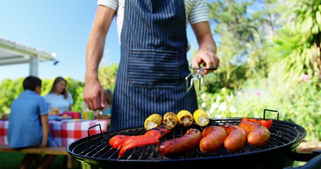 Man grilling a variety of meats and vegetables on a barbecue grill in a sunny backyard. In the background, a family sits at a picnic table featuring a checkered tablecloth. Ideal for use in articles about family gatherings, BBQ recipes, summer activities, or outdoor cooking tips.