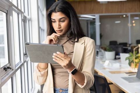Indian businesswoman reviewing tablet while standing at modern open-plan office window