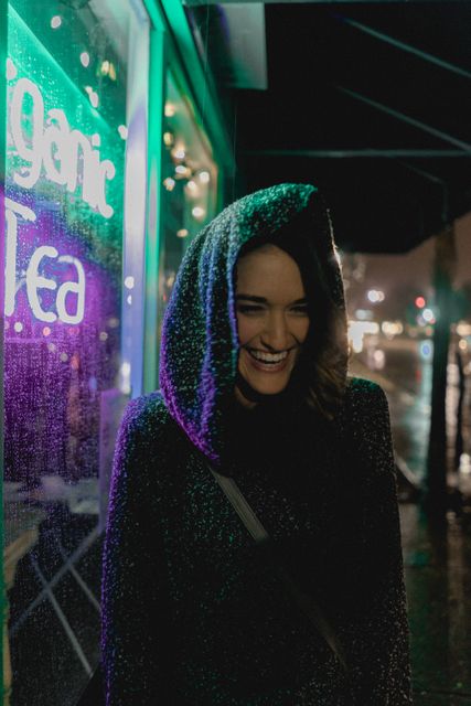 Smiling woman wearing a hoodie, standing on a rainy night under colorful lights reflecting on wet sidewalk. Background includes neon sign and blurred street lights creating a vibrant urban setting. Ideal for themes related to city life, youthful energy, nightlife, and cozy rainy day moments.