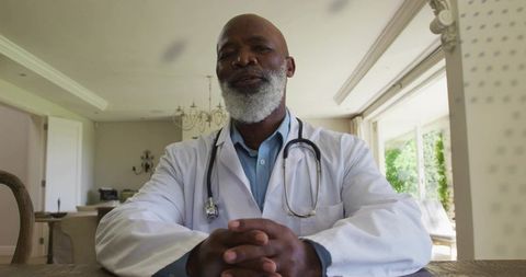 Senior doctor sitting at home table wearing white coat and stethoscope folding hands