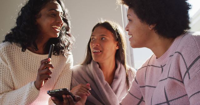 Three diverse women are sharing a light-hearted moment together while using a smartphone indoors. Perfect for illustrating themes of friendship, diversity, casual conversation, and bonding in a relaxed, comfortable setting.