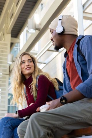 Young diverse students chatting on campus bench holding coffee and wearing headphones
