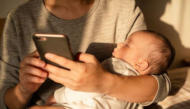 Caregiver holding sleeping newborn while scrolling smartphone in warm sunlit living room