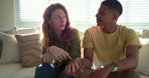 Sunlit diverse couple holding hands on couch sharing intimate conversation with smartwatch