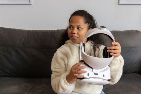Girl holding white vr headset on couch wearing cozy fleece and jeans exploring virtual reality