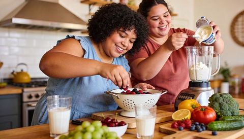 Joyful friends preparing berry smoothie together pouring yogurt into blender in kitchen