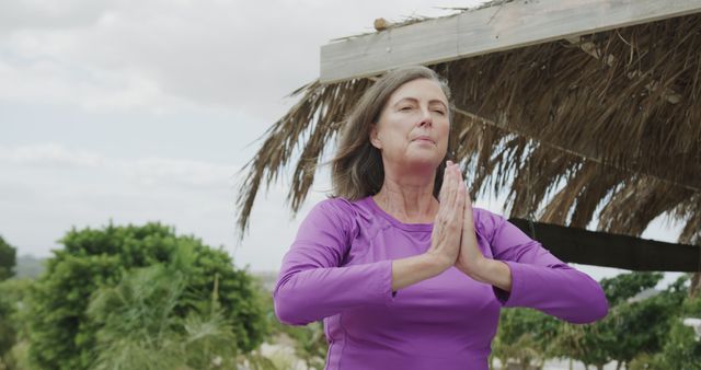 Mature woman practicing yoga by bringing hands together in prayer pose outdoors under open sky, surrounded by greenery. Gentle and peaceful expression on her face, indicating relaxation and mindfulness. Ideal for use in health and wellness promotions, mindfulness and meditation guides, fitness and lifestyle blogs, and advertisements focusing on healthy living and outdoor activities.