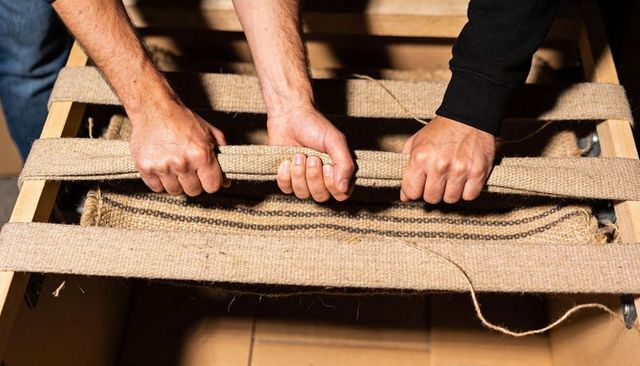 Two hands pulling jute webbing on wooden upholstery frame repair and furniture restoration