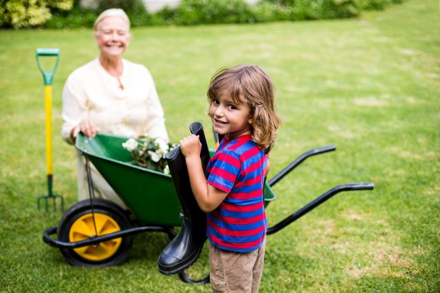 Boy holding wellington boots while standing next to his grandmother in a garden. Grandmother is sitting in a wheelbarrow with gardening tools nearby. Perfect for themes related to family bonding, outdoor activities, gardening, and summer fun.
