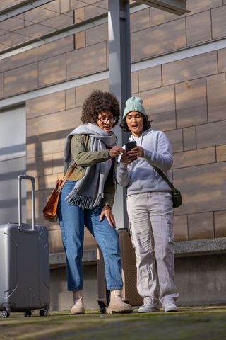 Female friends checking smartphone at transit stop with rolling luggage for urban travel