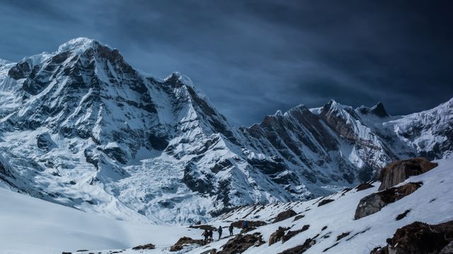 Several alpine climbers trekking snowy trails of a majestic mountain during twilight. The scene captures a sense of adventure, determination, and natural beauty with towering snow-covered peaks under darkening skies. Ideal for travel brochures, adventure magazines, advertisements for outdoor gear, and inspirational posters emphasizing challenge and perseverance.