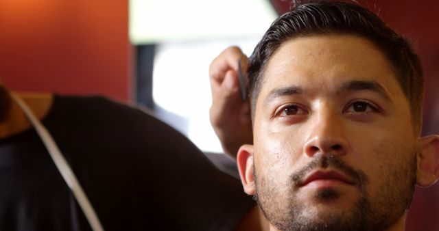 A young Asian man is getting a haircut from a barber, with copy space. His expression is contemplative as he looks off into the distance, considering his new style.