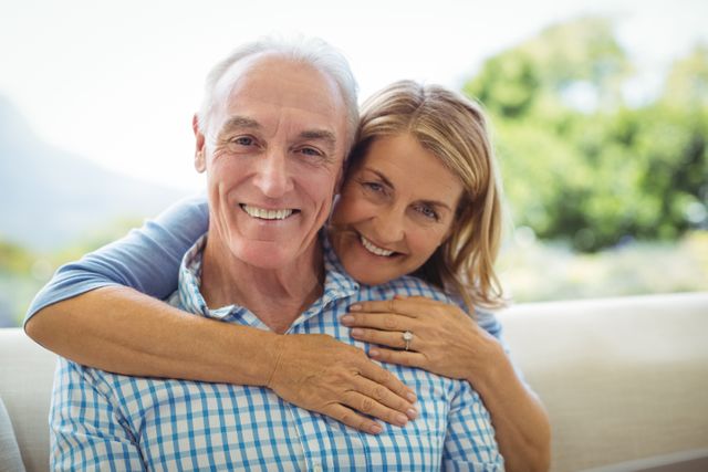 Senior couple smiling and embracing in a cozy living room. Perfect for use in advertisements, articles, and promotions related to senior living, retirement, family life, and relationships. Ideal for illustrating themes of love, happiness, and togetherness in later life.