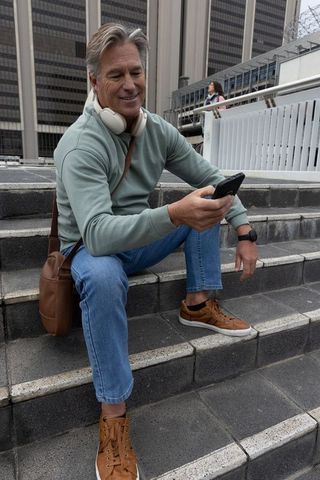 Senior man sitting on city steps checking smartphone with headphones and crossbody bag