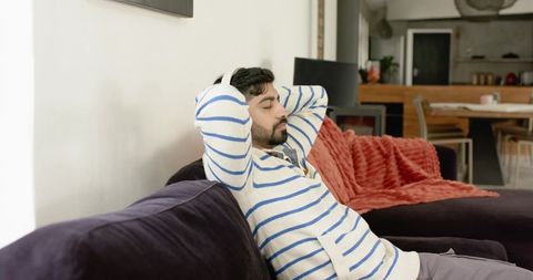 Indian man reclining on dark velvet sofa with headphones wearing striped sweater in cozy modern home