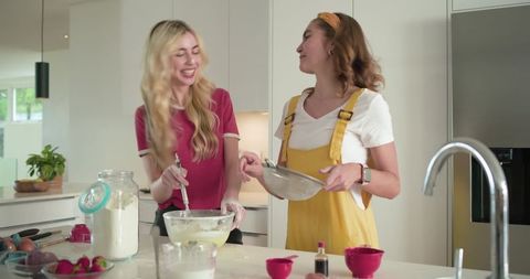 Two friends baking together in bright modern kitchen mixing batter and laughing