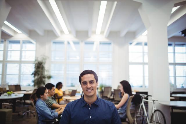 A man in a blue shirt smiles confidently at the camera while his colleagues are working in the background in a modern office setting. This can be used for business presentations, websites, brochures, or articles related to corporate environments, teamwork, and professional workspaces.