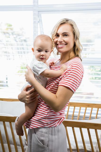 Smiling mother holding her baby boy in a bright room with natural light coming through the window. Ideal for use in parenting blogs, family-oriented advertisements, and articles about motherhood and child care.