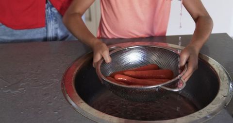 Child and parent draining hot dogs in stainless steel colander over round kitchen sink