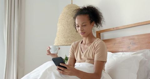 Relaxed Woman with Mug Scrolling Smartphone in Minimalist Bedroom