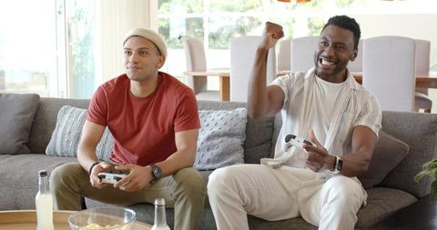 Friends playing video games on couch, one cheering after win in modern bright living room