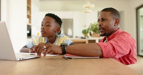 African american father tutoring son at kitchen table using laptop, notebook for homework