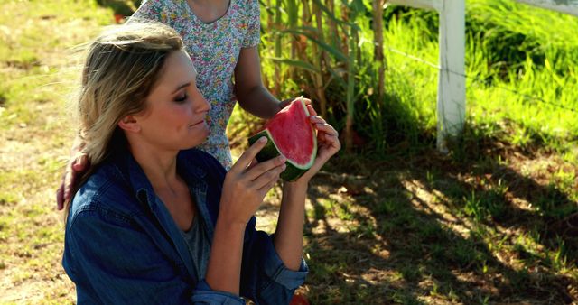 A young Caucasian woman enjoys a slice of watermelon outdoors, with a child partially visible in the background, with copy space. Moments like these capture the simple pleasures of summer and family time.