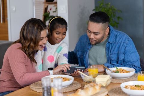 Family sharing smartphone at dining table while enjoying casual pasta meal and fresh bread
