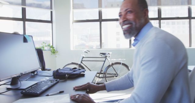 Smiling man sitting at a desk in a modern office setting with a computer monitor and a window letting in natural light. Ideal for use in business, workplace productivity, modern office environments, and positive work culture themes.