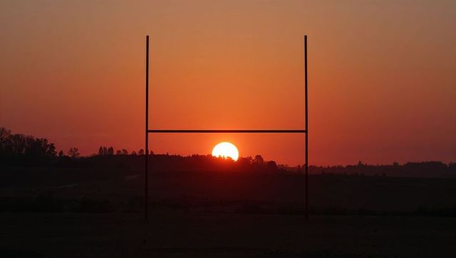 Sun setting framed by goalposts over rolling rural hills at vivid orange dusk