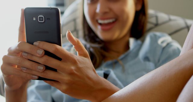 A young woman relaxes at home and uses her smartphone, smiling at what she sees on the screen. This stock image is perfect for illustrating concepts of modern communication, technology use, and relaxation. Ideal for articles, blogs, social media posts, and advertisements related to technology, daily life, or home decor.