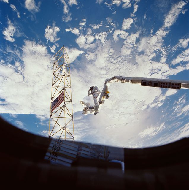 Astronauts Jerry L. Ross (right) and Sherwood C. (Woody) Spring (left) share a foot restraint as they survey the assembled ACCESS components after a lengthy extravehicular activity. Both men salute the American flag placed on the assembled ACCESS tower. Stowed EASE pieces are reflected in the window through which the photo was taken.