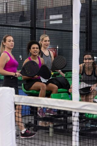 Multicultural women resting between padel matches holding paddles on indoor court bench