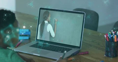 Child attending virtual classroom on laptop while teacher writing on chalkboard screen
