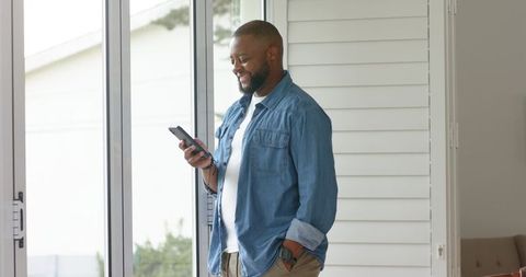Smiling man standing by sliding glass doors checking smartphone in modern home