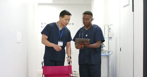 Asian and black male nurses consulting tablet in clinic hallway near red wheelchair
