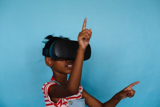 African american elementary schoolgirl gesturing while wearing vr glasses against blue background. unaltered, copy space, virtual reality simulator, playful, technology and school concept.