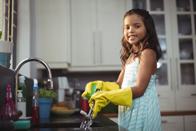 Young girl wearing yellow gloves washing dishes in a modern kitchen. She is smiling and appears happy while performing the chore. Ideal for use in articles about teaching children responsibility, family life, domestic chores, and home hygiene.