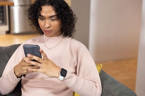 Non-binary person sitting on sofa using smartphone in apartment wearing pale pink sweater