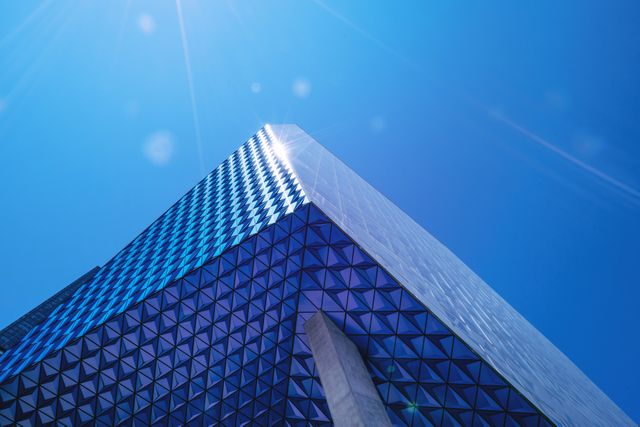 This image features a modern building with an abstract and geometric facade against a clear blue sky. The sun flares add a dynamic and futuristic touch to the scene. Ideal for use in content relating to modern architecture, innovation, urban development, futuristic designs, or real estate.