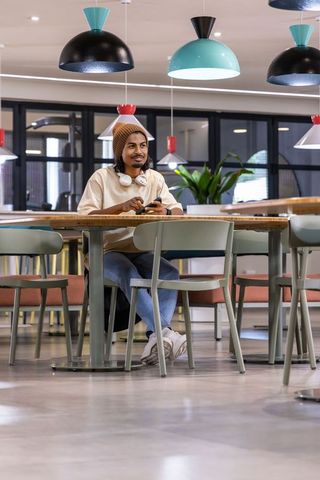 Indian man sitting in modern coworking space wearing headphones and holding smartphone