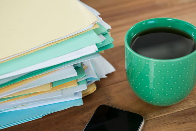 This image shows a close-up of a green coffee mug, a mobile phone, and a stack of files on a wooden table. It is ideal for illustrating concepts related to office work, productivity, organization, and business environments. It can be used in articles, blog posts, and presentations about office life, work habits, and organizational tips.