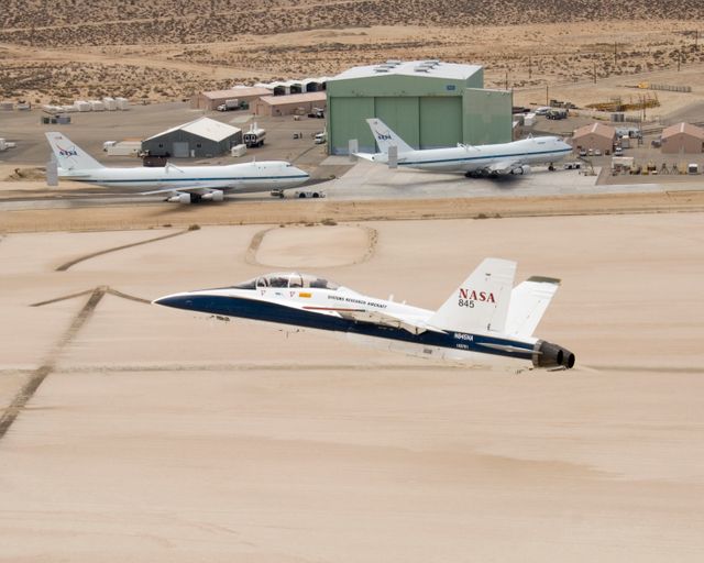NASA's two Boeing 747 Shuttle Carrier Aircraft form the backdrop as pilot Dick Ewers banks NASA F/A-18 #845 low over Rogers Dry Lake to end a research flight.