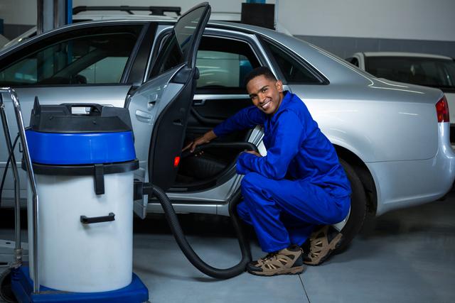 Mechanic in blue uniform vacuuming car interior in an auto repair shop. Ideal for illustrating automotive services, car maintenance, professional mechanics, and vehicle care. Can be used in advertisements for auto repair shops, car detailing services, and automotive maintenance tutorials.